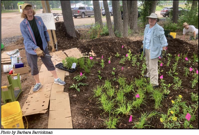 Two smiling women wearing hats and work clothes and gloves stand among over 100 new native plants marked by pink flags planted in a rain garden.